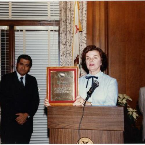 [Dianne Feinstein holding a plaque in City Hall with Consul General Romeo Arguelles on the left, 