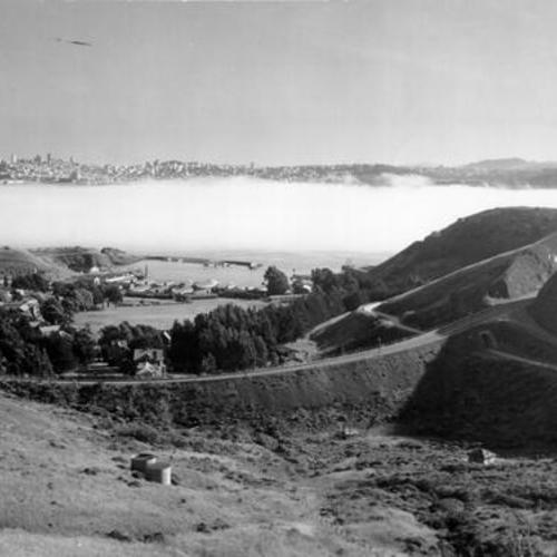 [View of San Francisco from the Marin hills, showing the Waldo approach to the Golden Gate Bridge and a layer of fog covering the bay]