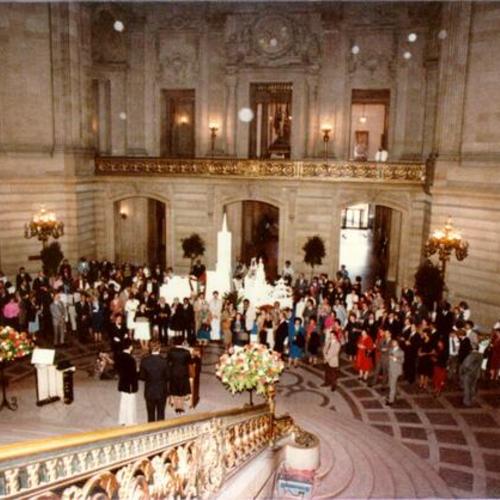 [Group of people gathering around a miniature structure of San Francisco while listening to a speaker in the Rotunda of City Hall]