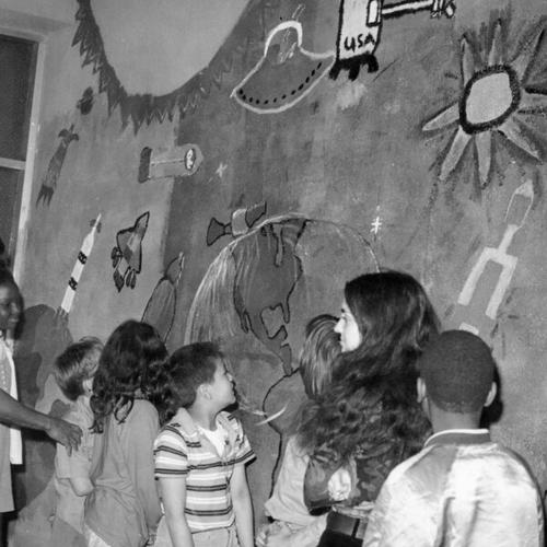 [Students and parent volunteer standing in front of space mural in the shed area of Sir Francis Drake Elementary School]