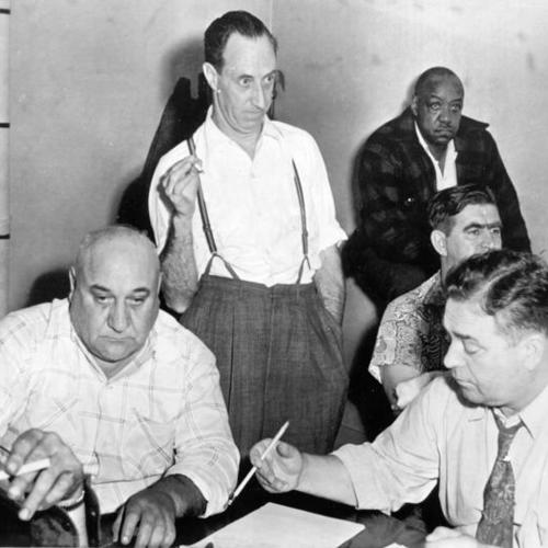[Harry Bridges (standing) shown at conference here today with Seattle maritime strike leaders. Jason Hopkins, local longshore president is seated (left) and secretary Jack Price (right)]