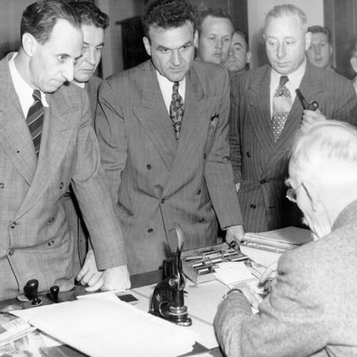 [U.S. Commissioner Francis St. J. Fox (seated in foreground) prepares papers on new bail for Harry Bridges (left) and his codefendants, J. R. Robertson and Henry Schmidt, after their sentencing in Federal Court]