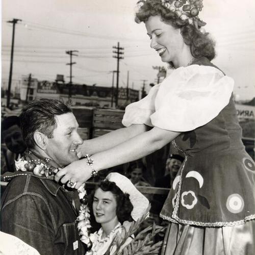 [Betty Jean Phillips, the "Queen of the Farmers' Market Fiesta," placing a wreath of garlic around the neck of apple grower Edward Holzreiter]