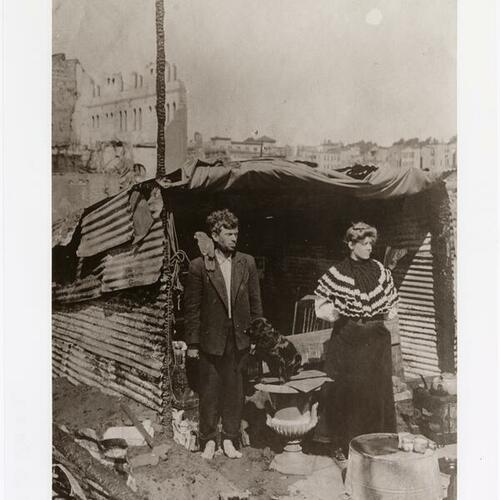Two people standing in front of makeshift shelter with one having a parrot on their shoulder and a dog