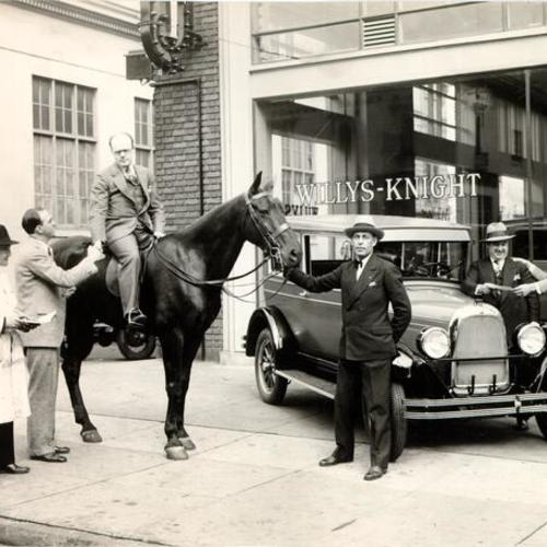 [J. H. Stephens (on horse) and Walter Packard (holding bit) in front of Willys-Knight automobile dealership]