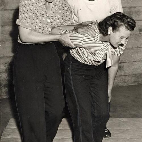 [Elizabeth M. Rickey puting an armlock on her classmate Florence Moodie while Officer Fred Fitzgerald checks on her methods]