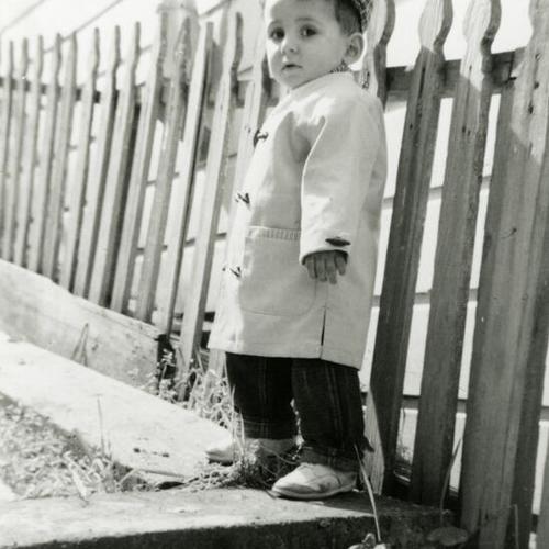 [Child with a cap and jacket next to a fence]