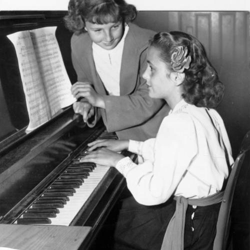 [Evangeline Powell playing piano while Georgia Ingmire turns the pages of the sheet music for her at Everett Junior High School]