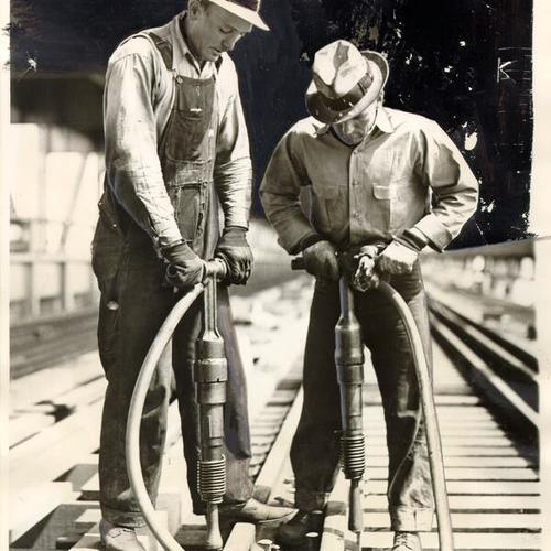 [Workers Harry Keene and Frank Hauslik driving spikes with pneumatic hammers during construction of an electric railway system across the San Francisco-Oakland Bay Bridge]