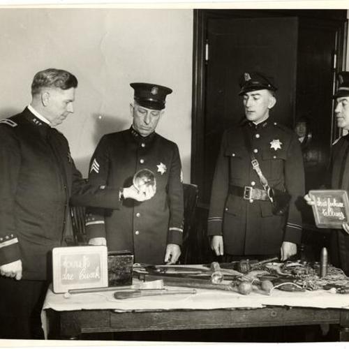 [Chief W.J. Quinn inspecting an object; Sarg. McGee, Henry M. Schutzer, Capt. Thomas M. Hoertkorn standing behind a table covered with different weapons]