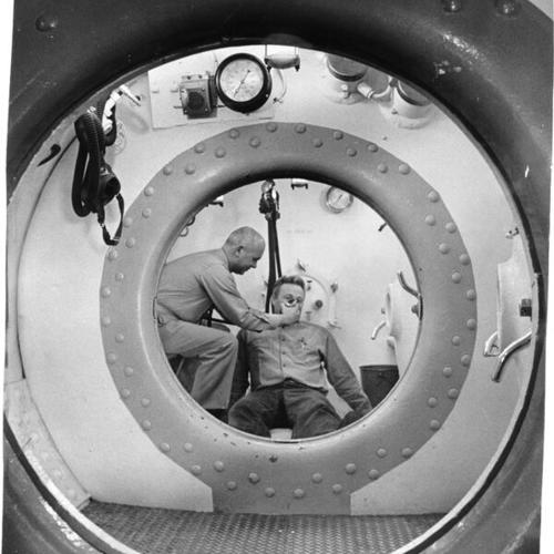 [Lieutenant Benjamin Dew administering treatment to Diver I/C John McAllister in a decompression chamber at Hunters Point Naval Shipyard]