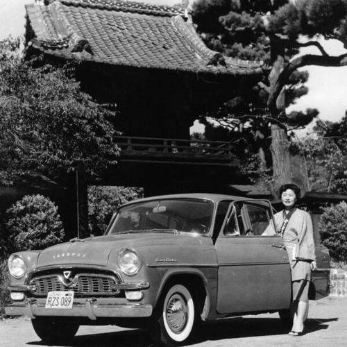 [Kim Furuya stands beside the Toyopet Crown Custom in front of the entrance to the Japanese Tea Garden]
