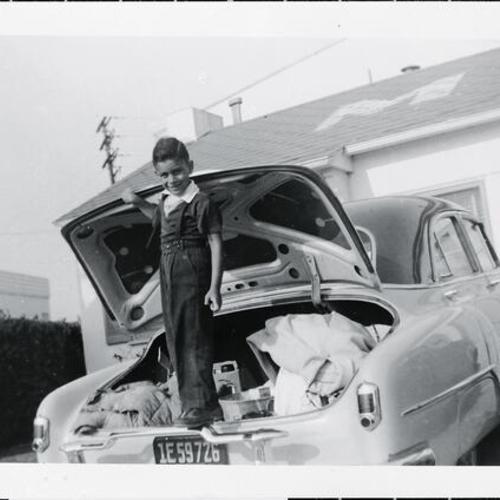 [Portrait of little boy standing on bumper and trunk of Chevy car]