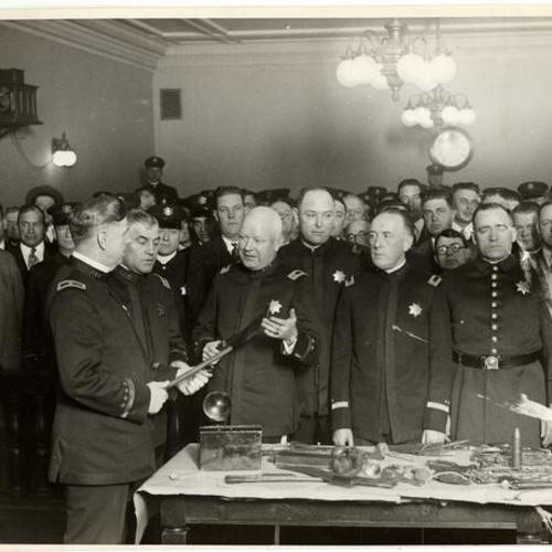 [Chief W.J. Quinn inspecting a weapon; Capt. Hoertkorn, Capt. McGowan, Lt. M. Mitchell, Lt. Pat Murray, Off. Pat Watch, Off. Sam Miller standing behind a table covered with different objects inside a crowded courtroom]