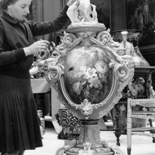 [Thelma Hamblin, staff member at the De Young Museum, dusting an elaborate burnished and enameled urn in preparation for a public auction.]