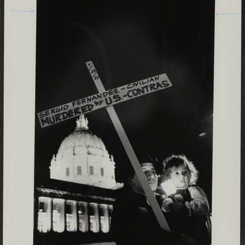 Mourner with child holding candle and cross at memorial in front of Federal Building commemorating Americans slain by contras