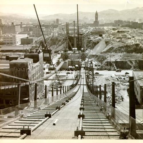 [View of the San Francisco-Oakland Bay Bridge while under construction showing suspension bridge catwalk extending from Rincon Hill]