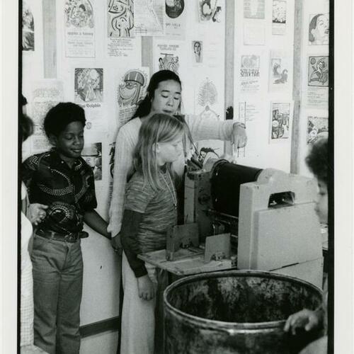 [A woman assists two children with a printing machine at South of Market Cultural Center Blind Workshop]