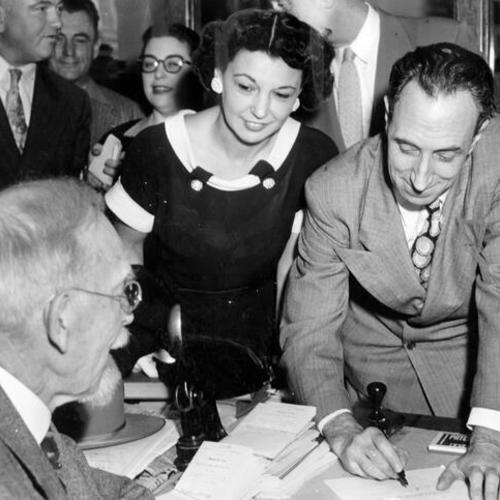 [Harry Bridges signing one of the documents in connection with his release on bail, as U.S. Commissioner Francis St. J. Fox looks on and Bridges' wife, Nancy, smiles her approval]