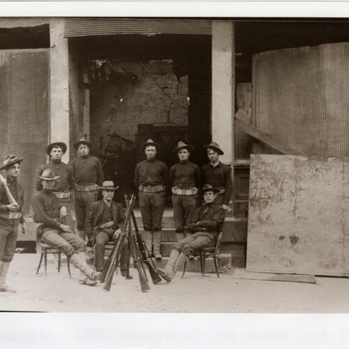 Soldiers gather in front of damaged building with riffles standing in the center