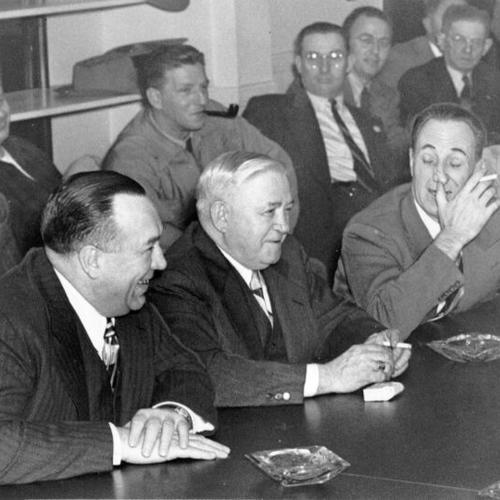 [Harry Bridges, head of the I. L. W. U., at right, scratches his nose reflectively as negotiations looking to end of the dock strike begins. At left are R. J. Thomas and Allan Haywood, of the National C.I.O.]