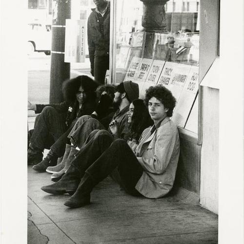 [Group of people sitting on a sidewalk in the Tenderloin District, near Market Street]