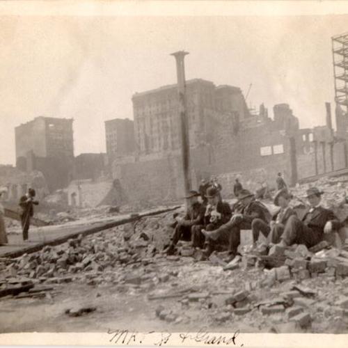 [Group of men sitting among ruins at Grant Avenue and Market Street, St. Francis Hotel in background]