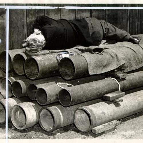 [Man lying on a bed of pipes on 'Skid Road']