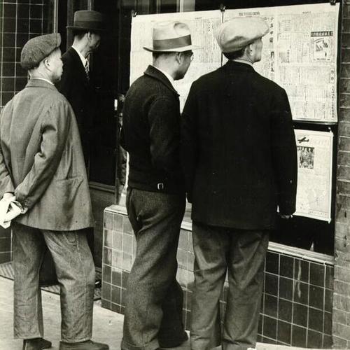[Group of men reading from newspaper notices outside The Young China, in Chinatown]