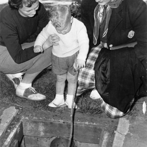 [Mary Kneis, Janice Sack and a small child look at a potentially dangerous open catch basin near an unidentified lake in Golden Gate Park]