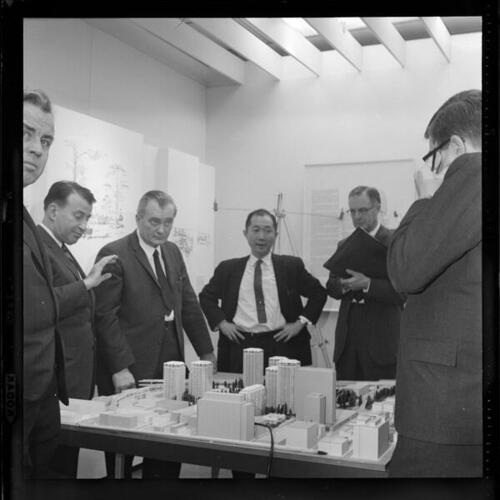 [A group of men discuss plans around an architectural model of downtown]