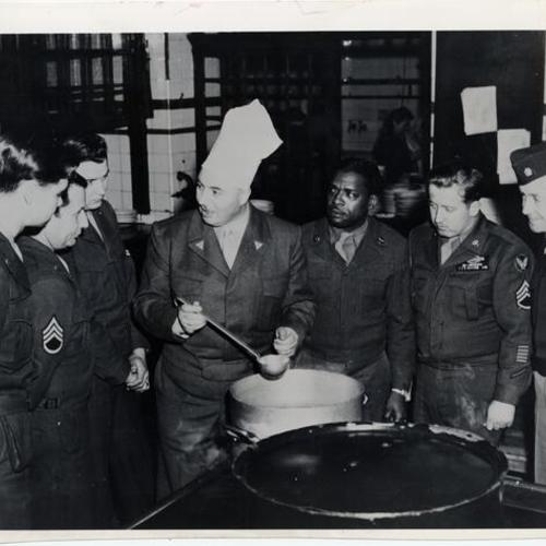 [George Mardikian (center) giving pointers on making soup to a group of U.S. Air Forces Mess Sergeants]