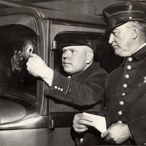 [Officer Joseph McVeigh inside a car examining a hole in the windscreen while Officer Jack Mangan observes from outside the car]