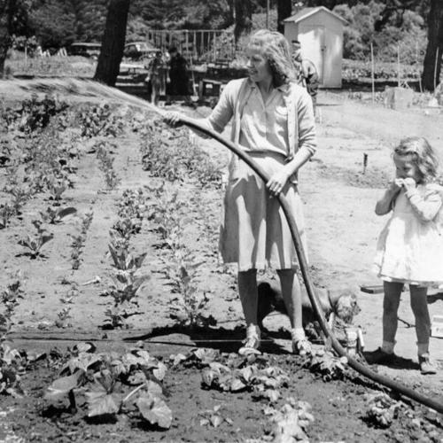 [Mrs. Rodney George irrigates her garden, with her daughter Eleanor looking on]