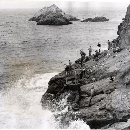 [Group of people standing on rocks fishing beneath the Cliff House]