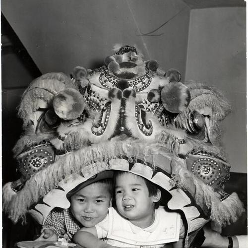 [Two young children playing under a lion's mask]
