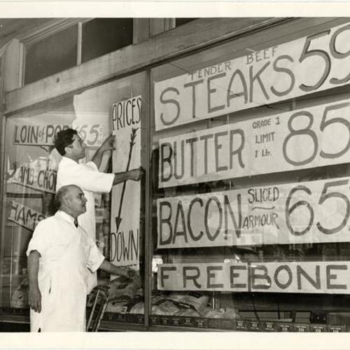 [Two men putting up a sign on the window of a butcher shop in Visitacion Valley]