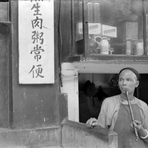 [Man with long opium pipe, in doorway. Goods for sale in shop window above him. Chinese sign on left]
