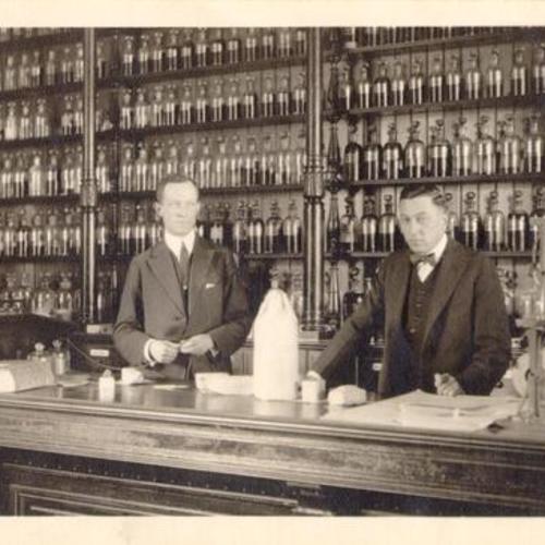 [Two men standing behind the counter at Broemmel's Pharmacy]