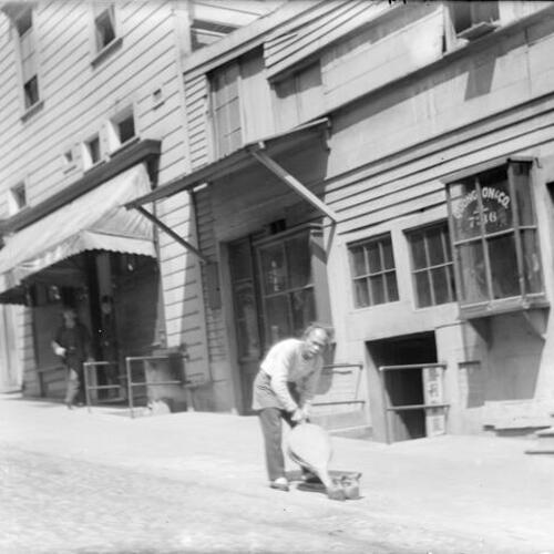 [Man on sidewalk using bellows to fire-up charcoal to beat two irons;