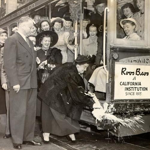 [Mrs. Robert A. Roos smashing a bottle of champagne at a ceremony marking the adoption of a cable car by Roos Bros. store]