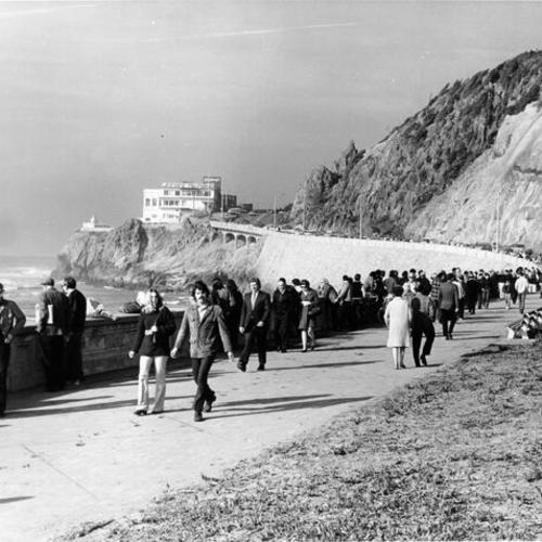 [People walking along a sidewalk at Ocean Beach, near the Cliff House]