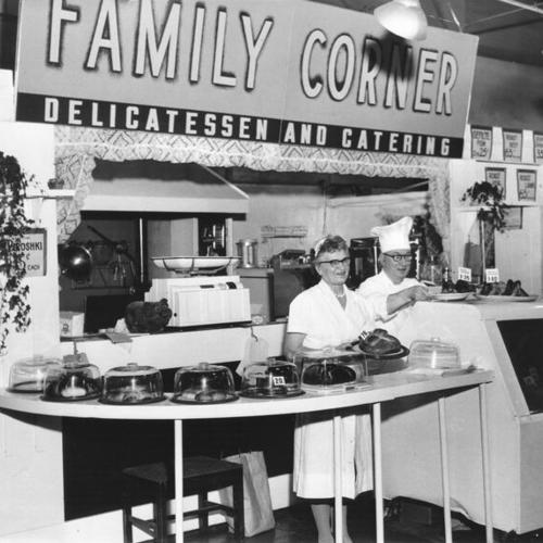 [Two employees standing behind the counter of the Family Corner delicatessen and catering counter at the Crystal Palace Market]