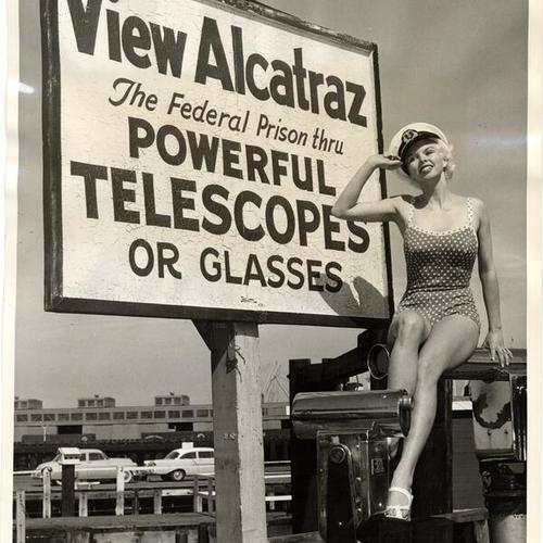 [Sue Boomer sitting next to a sign advertising telescopic views of Alcatraz]