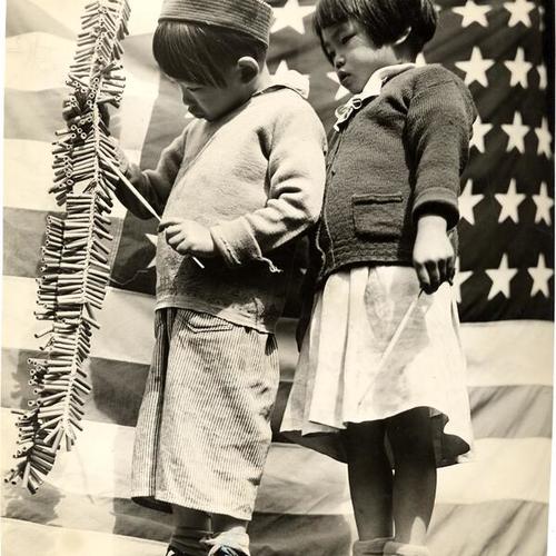 [Richard Mark and Thelma Lee standing in front of the american flag with a string of fire crackers]