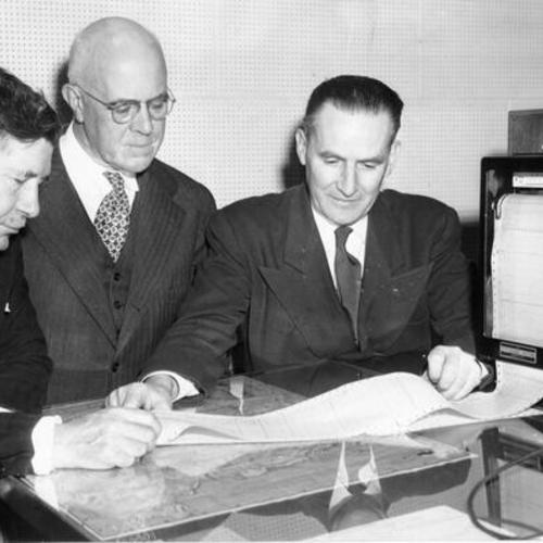 [Three Public Utilities officials looking at a graph, showing information about streetcar operation, at the Geary Street carbarn]