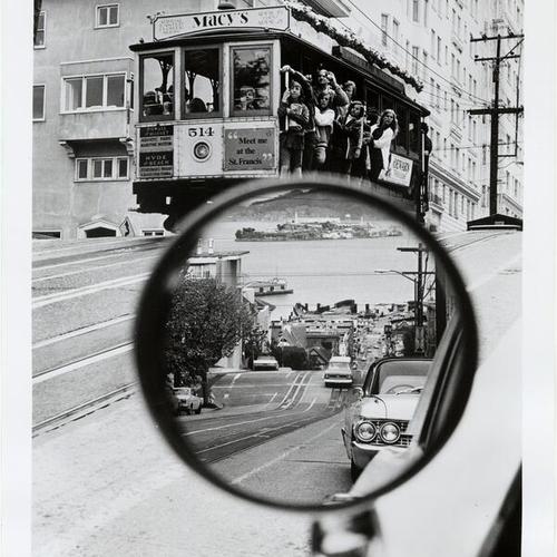 [View of cable car descending Hyde Street hill with its route reflected in the side-view mirror of a parked car]