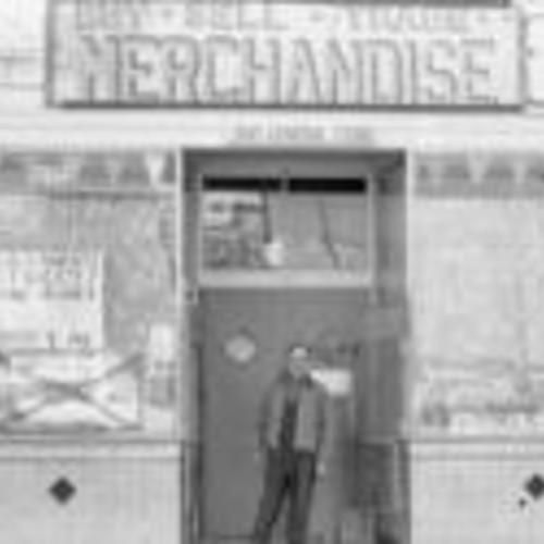 [Man (proprietor?) stands in doorway of Jim's General Merchandise,