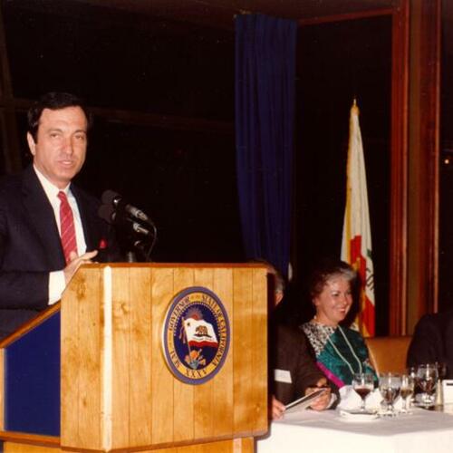 [Agnos speaking at podium with Shirley Temple Black and Governor George Deukmejian seated]