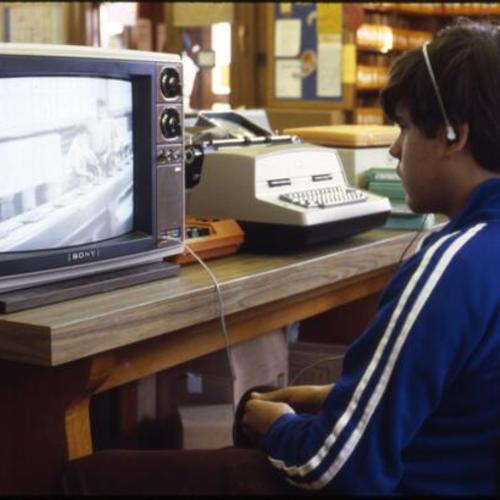 [A young man sits watching a video on a television next to a typewritter.]